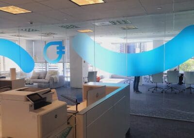 Modern office space featuring blue wave-like decals on transparent glass partitions, with workstations and a sitting area visible.